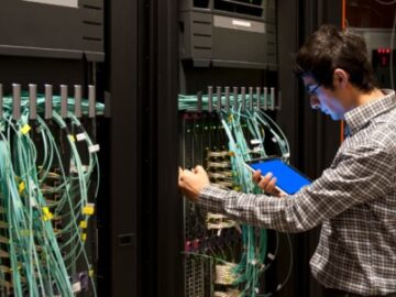 An IT professional with a tablet stands before a server rack, engaged in monitoring or managing IT equipment.