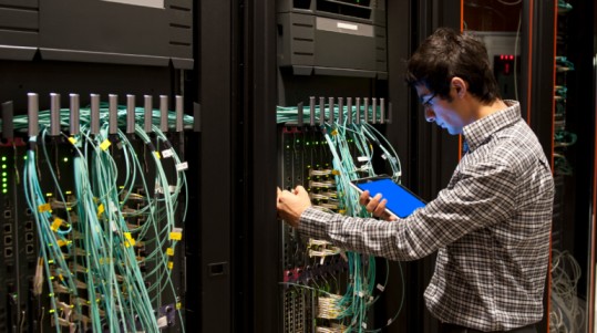 An IT professional with a tablet stands before a server rack, engaged in monitoring or managing IT equipment.